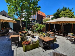 Een terras buiten met tafels en stoelen, omringd door groen en een decoratieve rotstuin.