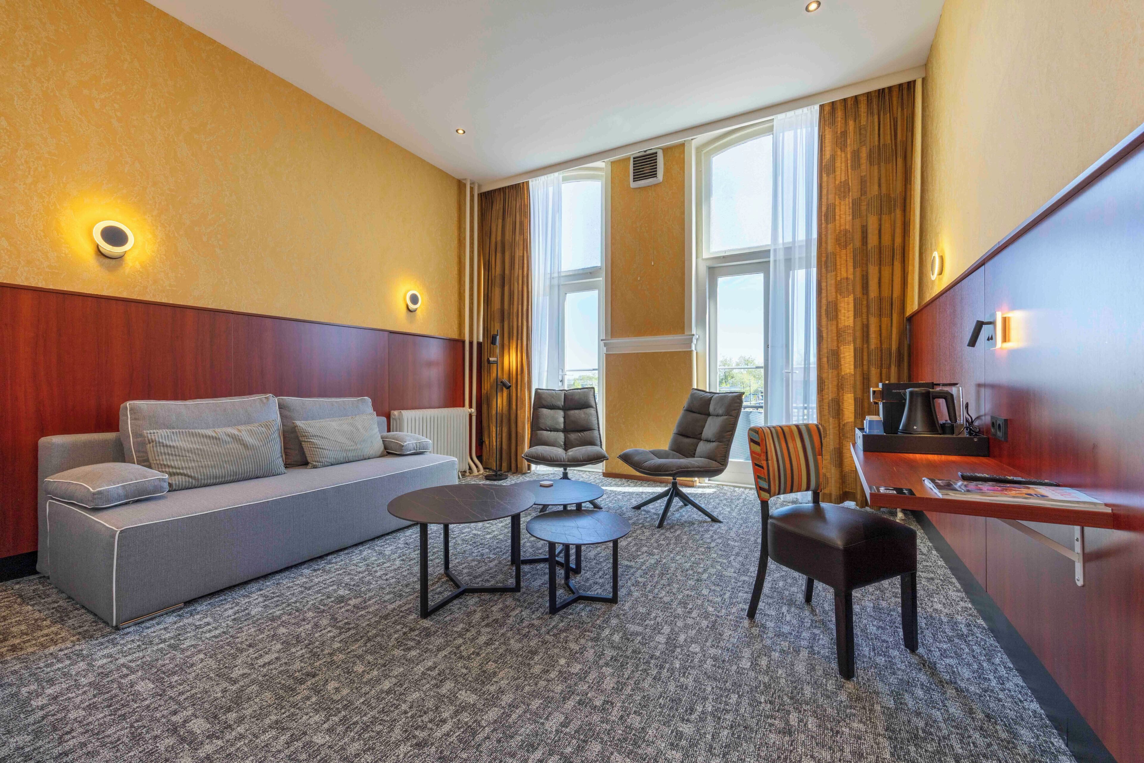 Habitación de hotel con sofá, dos sillones, una mesita, escritorio con cafetera y ventanas grandes con cortinas.