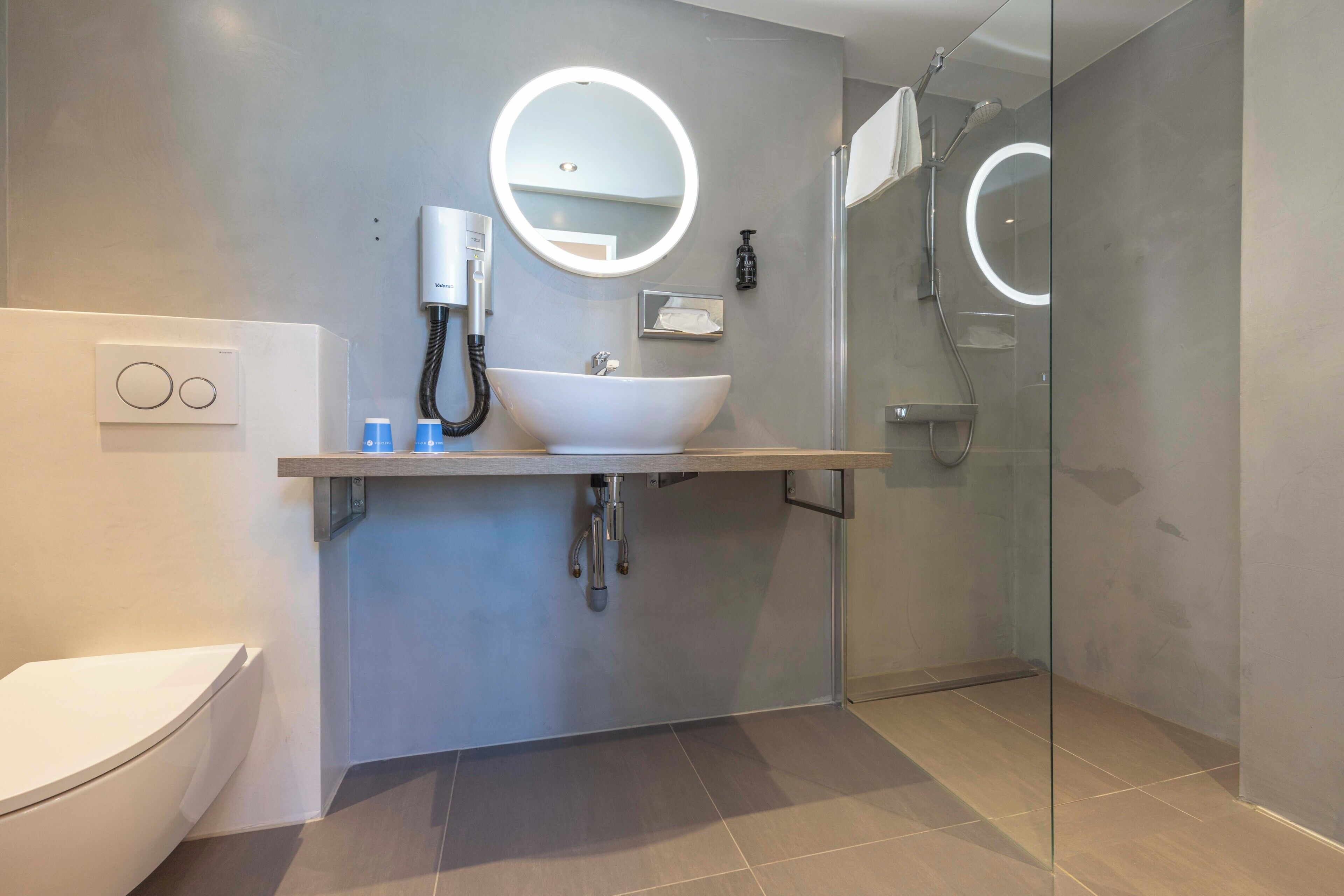 Ein modernes Badezimmer mit einem weißen Waschbecken, einem runden Spiegel, einer Handbrause und einer Duschkabine aus Glas.