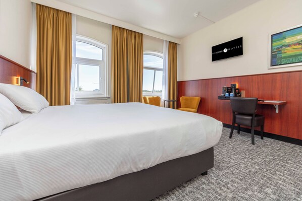 Quarto de hotel com uma cama grande, uma escrivaninha com cadeira, televisão e um quadro na parede.