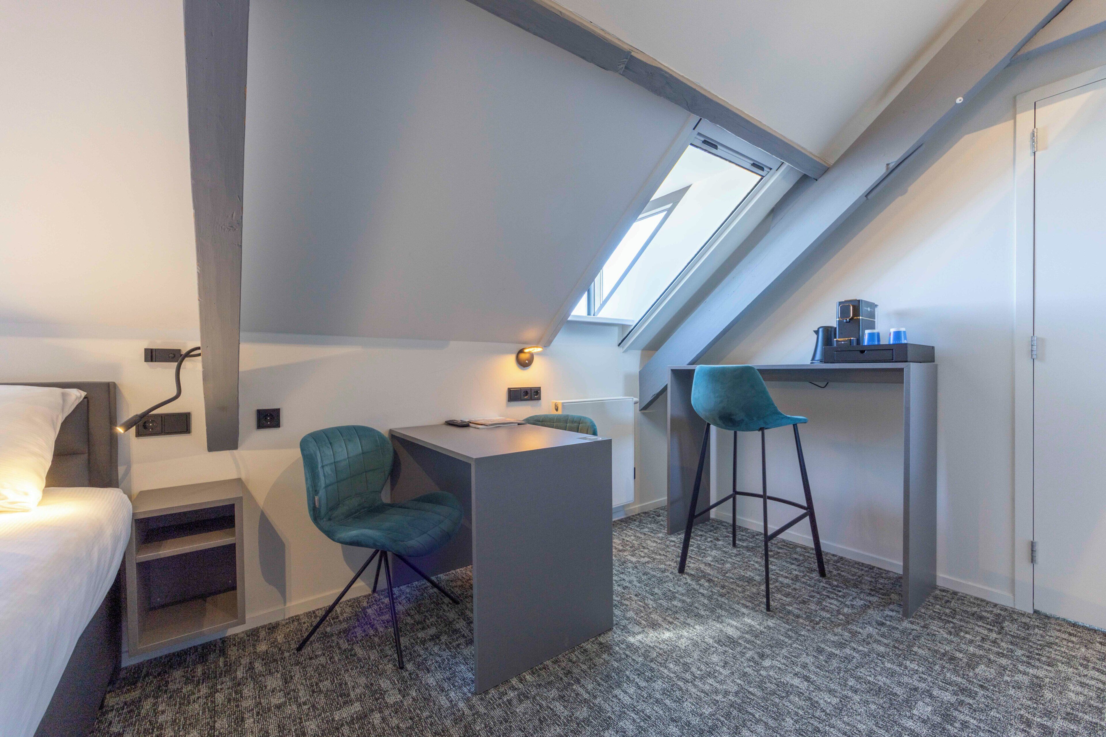 Een kleine kamer met een bed, een bureau, twee stoelen, een tafeltje en een dakraam.