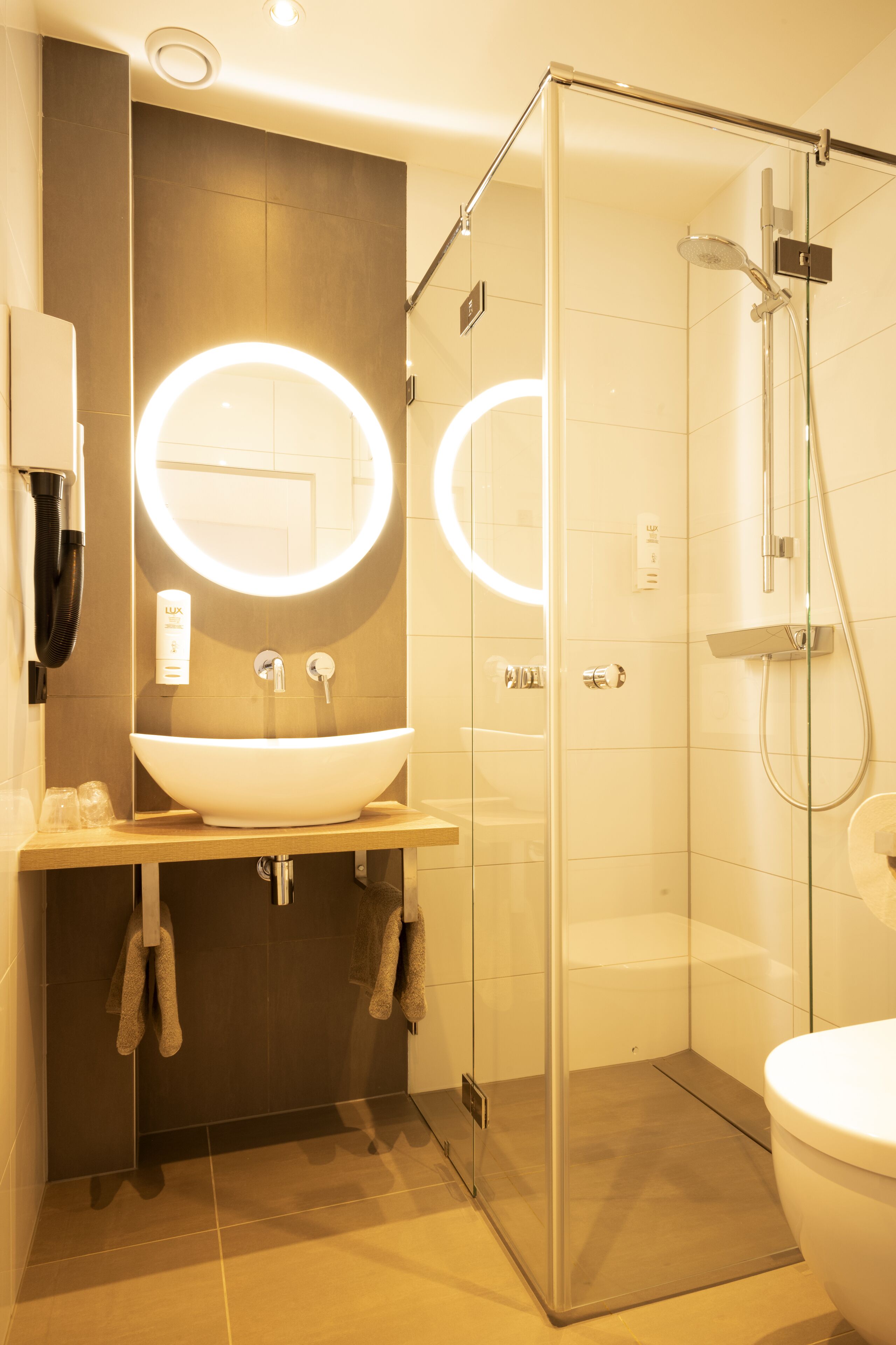 A modern bathroom with a glass-enclosed shower, a large oval sink, and dual illuminated mirrors.
