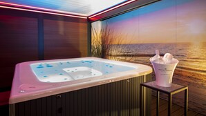 Un moderno espacio de spa con un jacuzzi, una pequeña mesa con una botella de champán y una vista panorámica del océano a través de amplias ventanas.