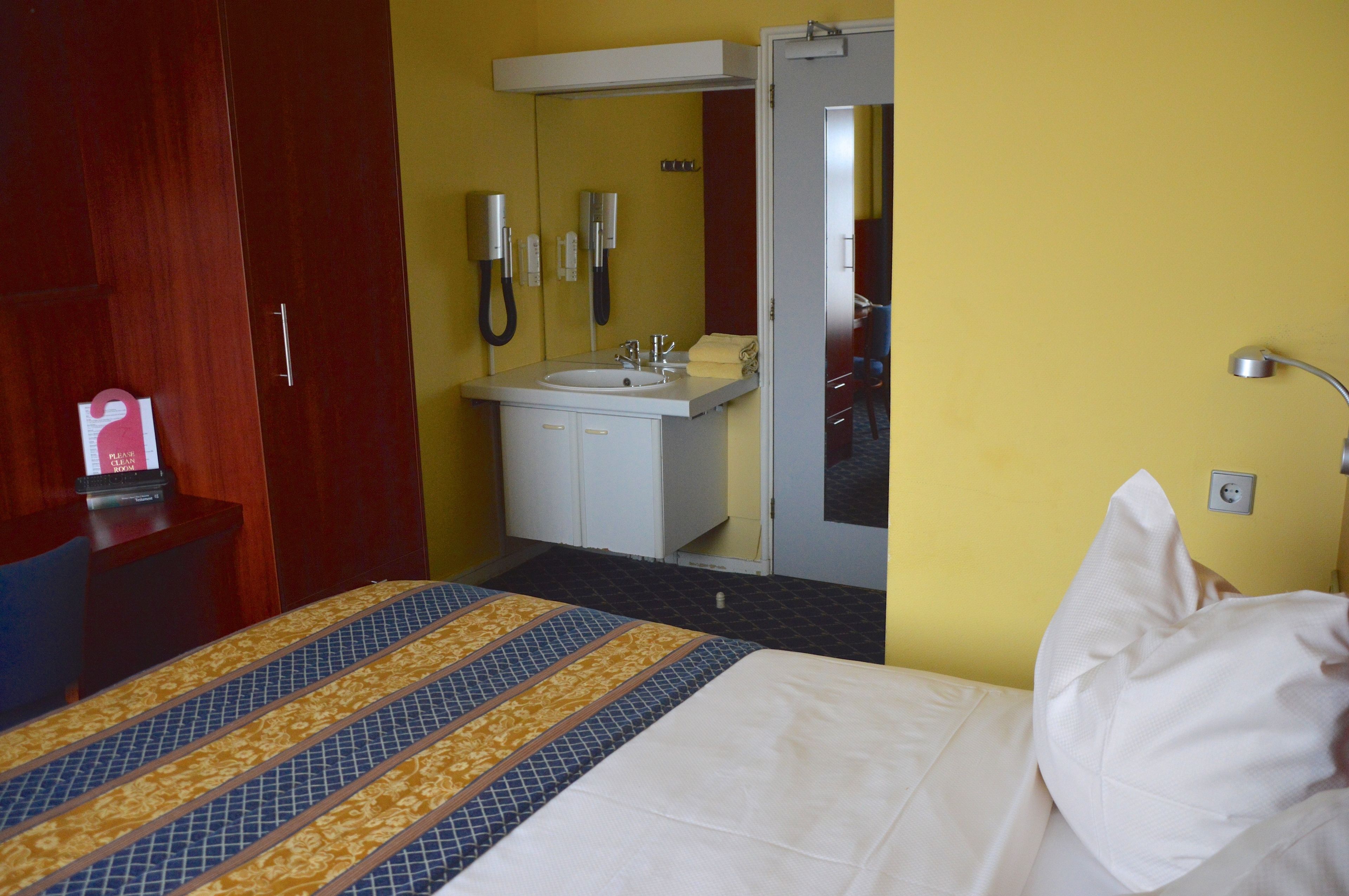 Una habitación de hotel con una cama, un baño con lavamanos y dos teléfonos, y un armario de madera.