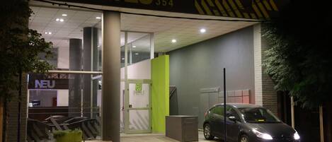Entrada de hotel con diseño moderno, que incluye fachada de vidrio, pared de acento verde y un coche estacionado afuera.