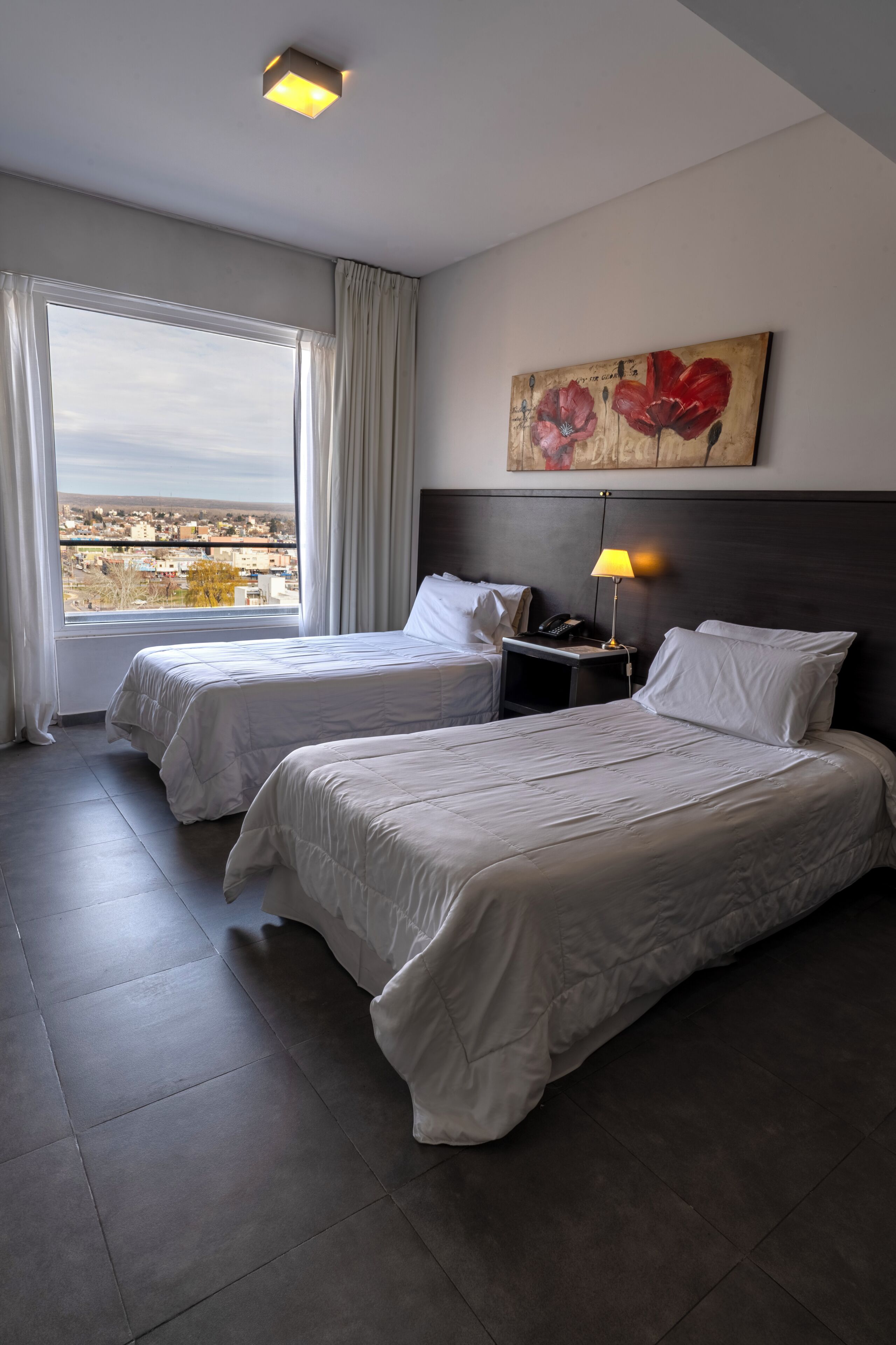 A hotel room with two beds, a nightstand, a lamp, a painting, and a view of the city.