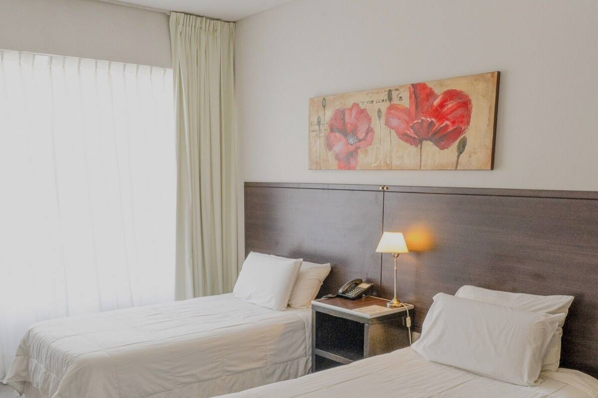 A hotel room with two beds, a nightstand with a lamp, and a painting of red flowers on the wall.