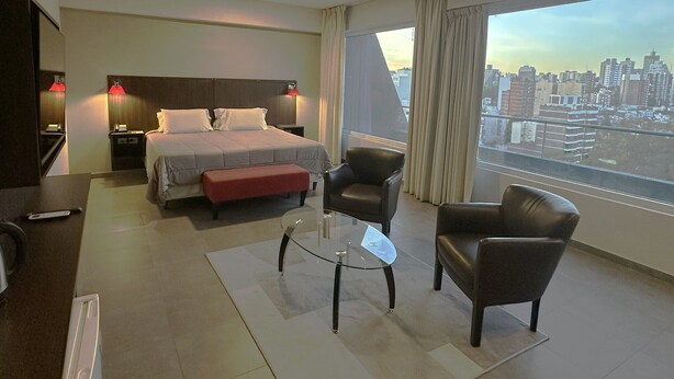 A hotel room with a bed, two chairs, a small table, and a city view. - Hotel Neu 354 (Neuquén)