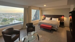 A hotel room with a large bed, a glass coffee table, two chairs, a red bench, and a city view through the window. - Hotel Neu 354 (Neuquén)