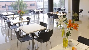 A modern dining area with multiple tables and chairs, large windows offering a view of buildings and trees, and decorative flowers on tables. - Hotel Neu 354 (Neuquén)