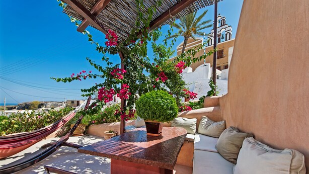 A sunlit outdoor seating area with a stone table, cushioned chairs, and a hammock. - Azalea Houses (Santorini)