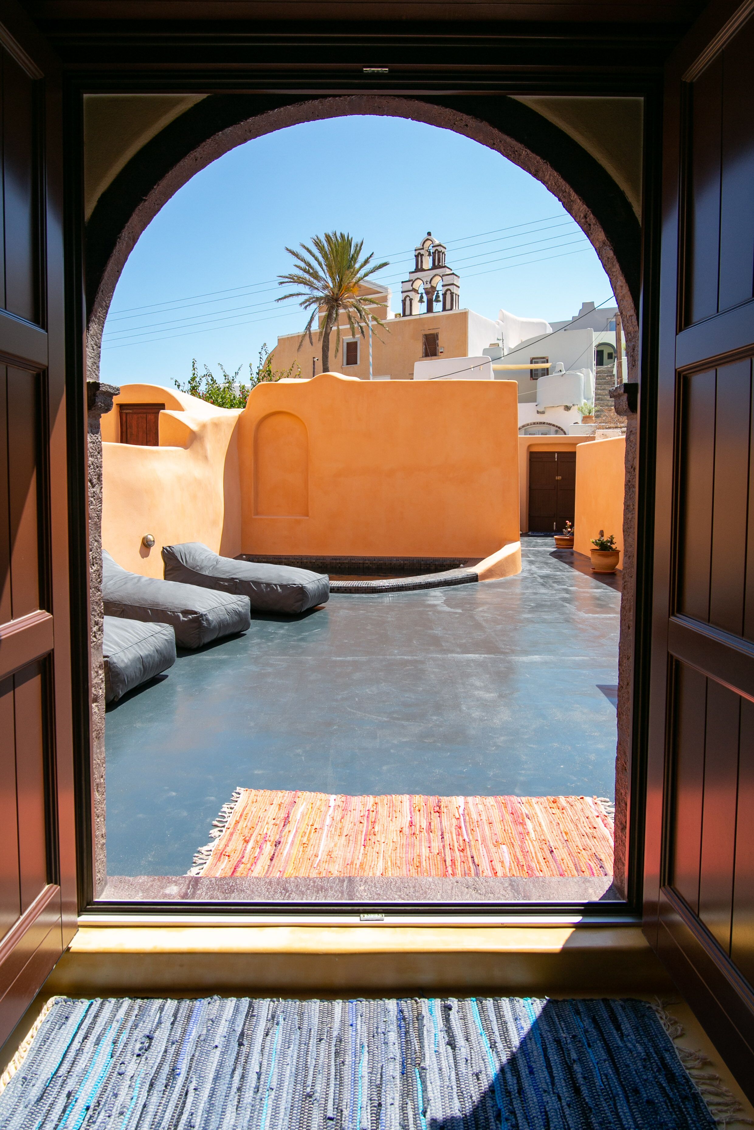 An arched doorway leading to a courtyard with a palm tree, a building with a bell tower, and a blue floor.