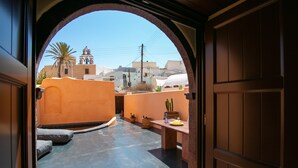 An open doorway leading to a courtyard with a view of a building with a bell tower, potted plants, and a tiled floor. - Azalea Houses (Santorini)