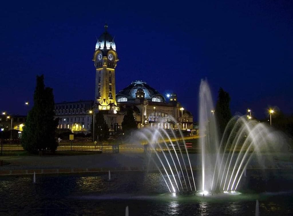 A night scene with a water fountain in the foreground and a building with a clock tower in the background.