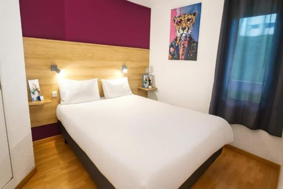 Habitación de hotel con cabecera de madera, cama grande con ropa blanca, mesita de noche con lámpara y un cuadro de un leopardo en la pared.