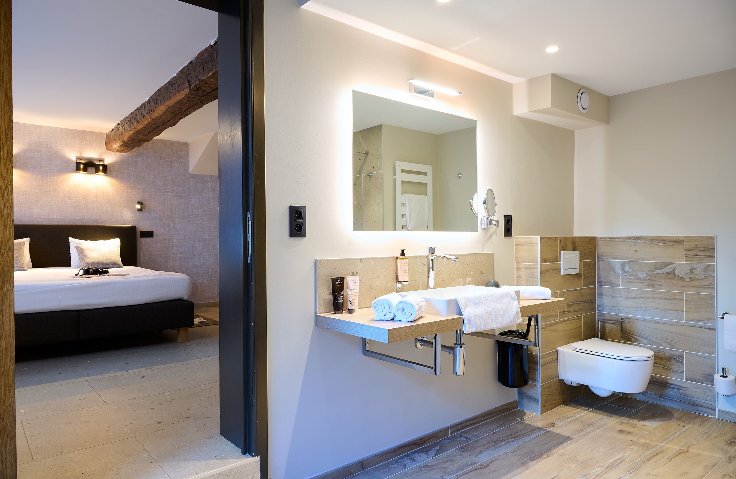 A modern bathroom with a large mirror, a sink, and a toilet. There is a bed with a headboard in the adjacent room.