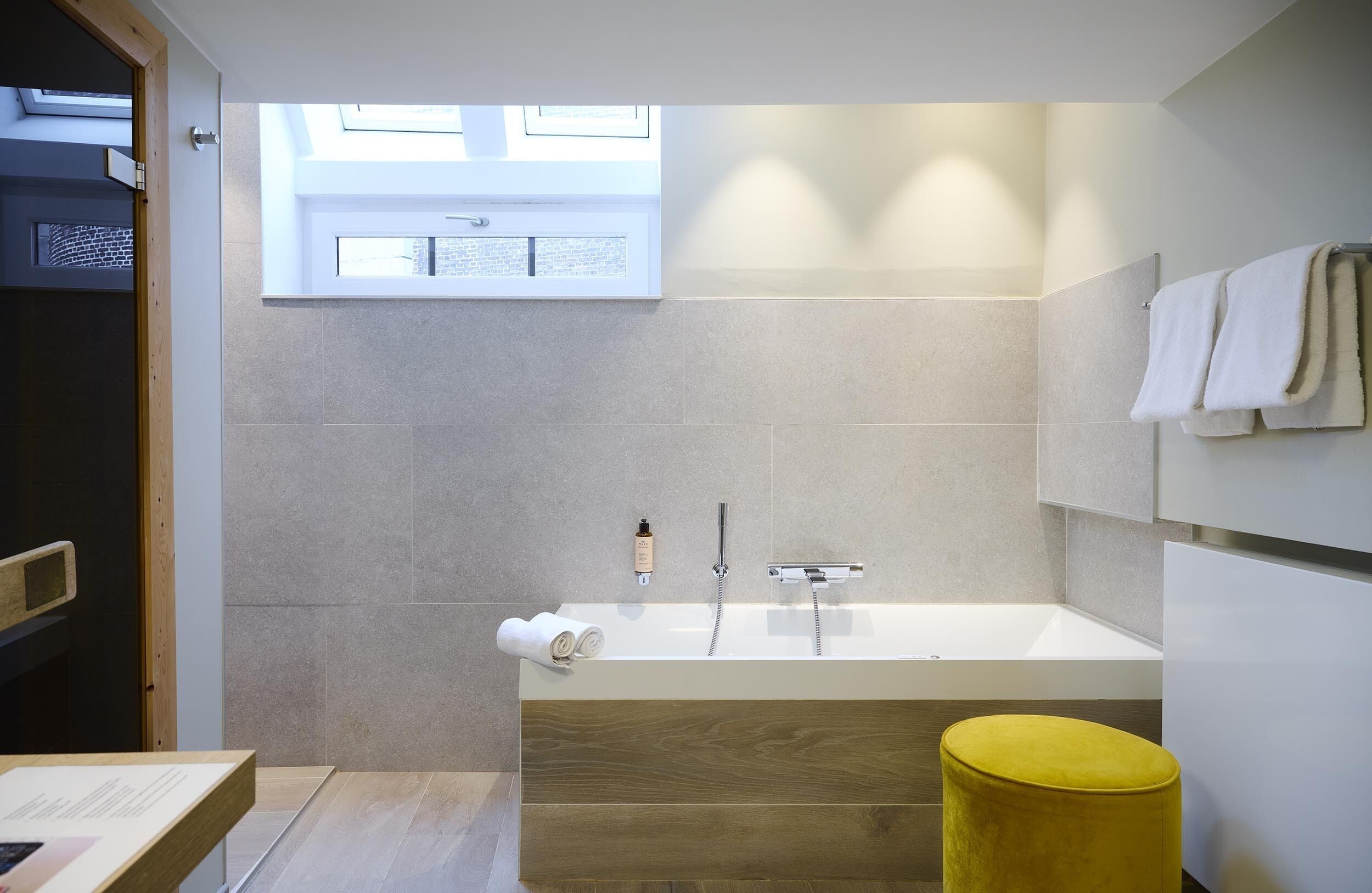 A modern bathroom with a large bathtub, a wooden bench, a yellow stool, and a window providing natural light.