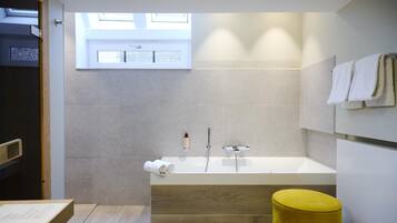 A modern bathroom with a large bathtub, a wooden bench, a yellow stool, and a window providing natural light.