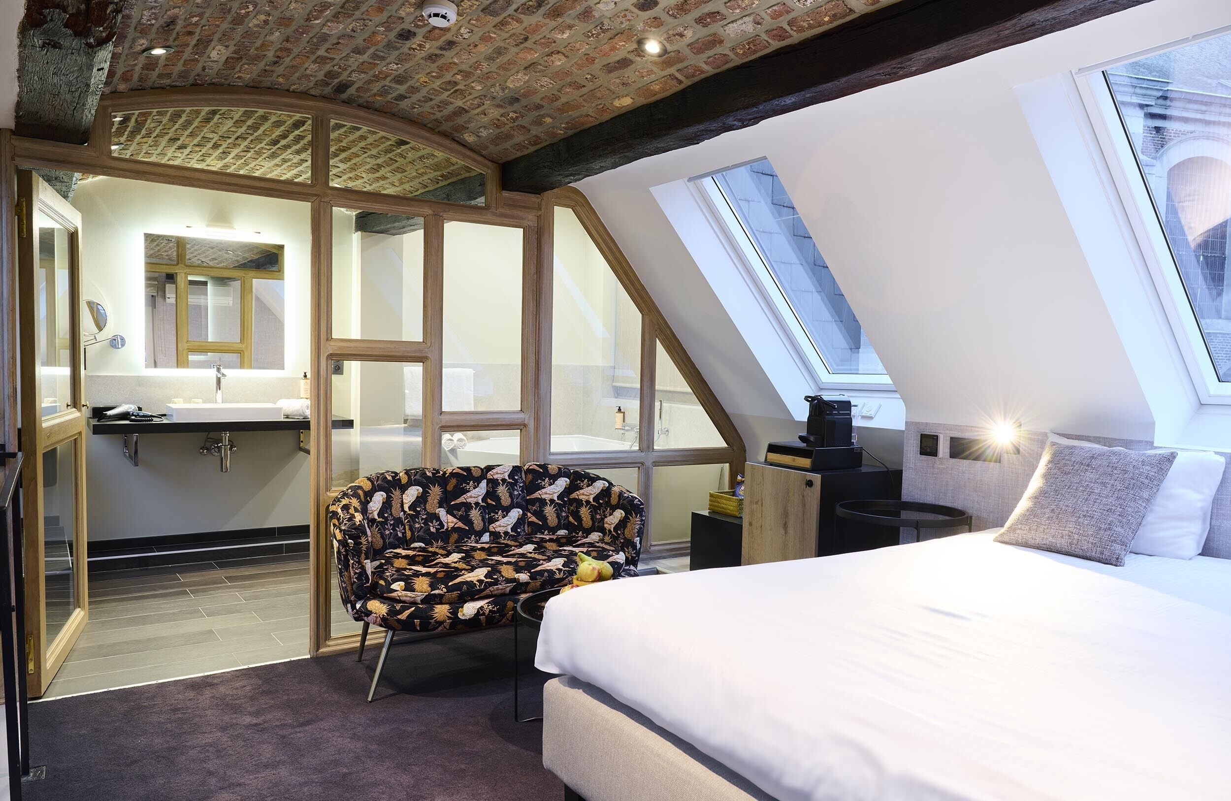 Een slaapkamer met dakraam, een bed, een stoel en een badkamer met spiegel en wastafel.