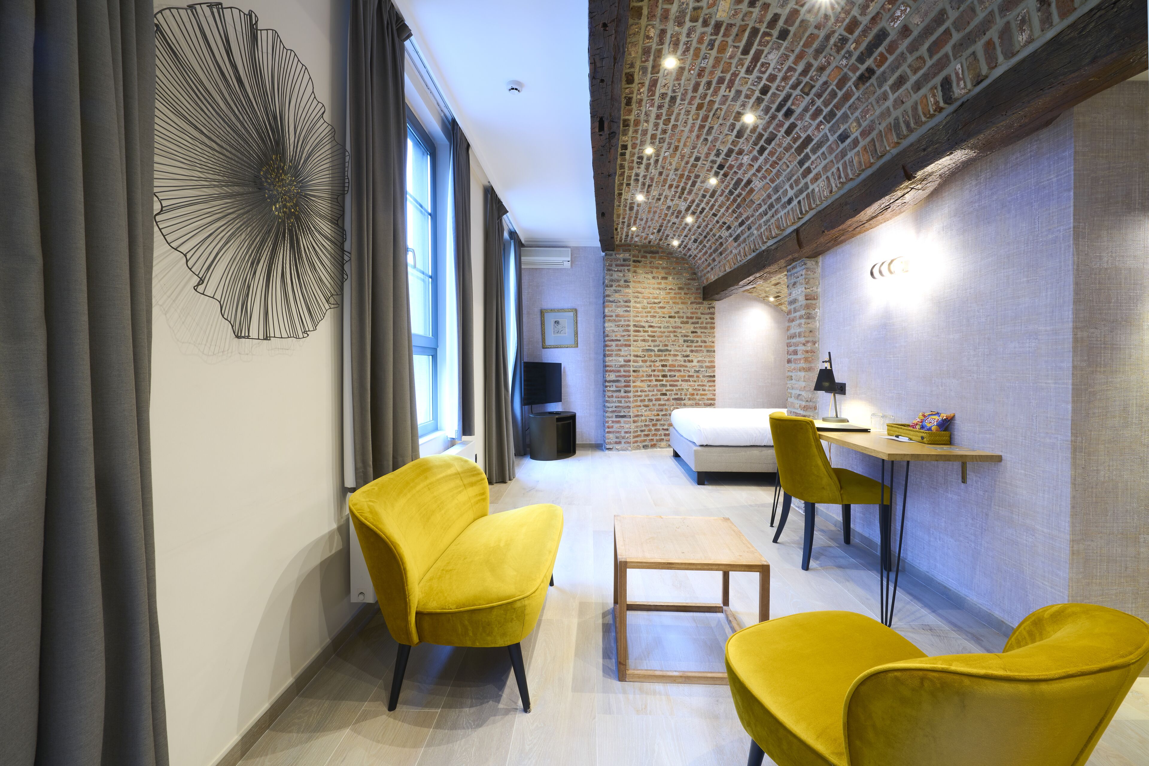 Una habitación de hotel moderna con pared de ladrillo, un ventanal grande, una cama, un escritorio y dos sillas amarillas.