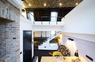 Un interior moderno con paredes de ladrillo visto, una escalera que lleva a una zona de altillo y una mesa de comedor con una lámpara.