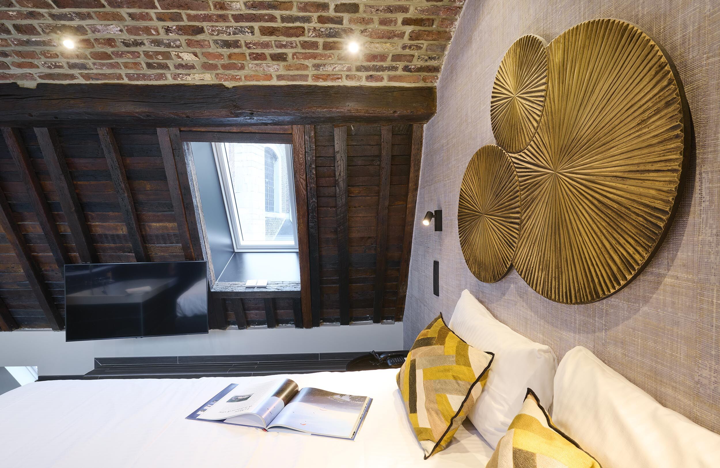 Een moderne hotelkamer met zichtbare bakstenen muren, een groot raam, een flatscreen televisie, een bed met gestreepte kussens en een decoratief kunstwerk aan de muur.