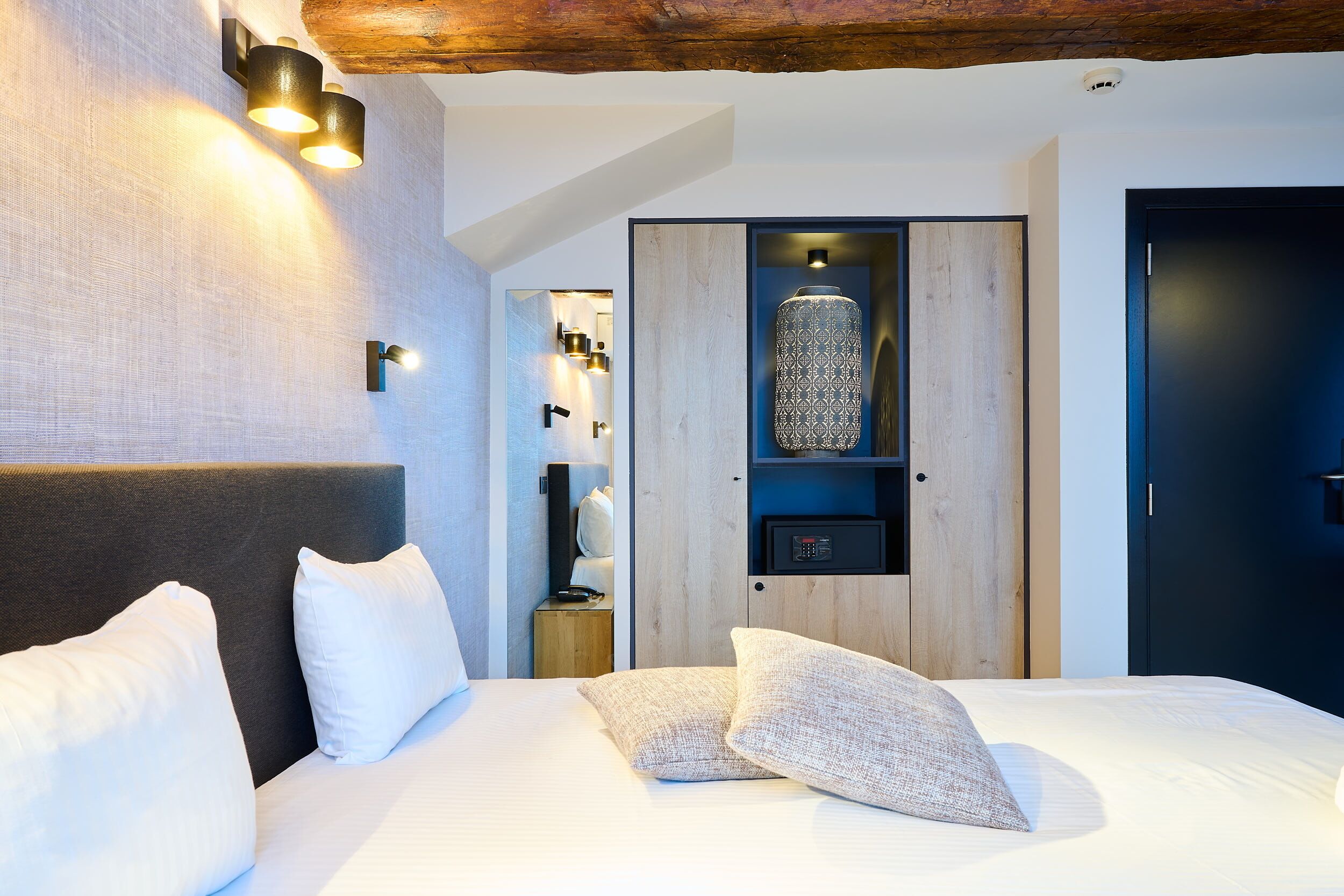 A modern hotel room with a large bed, a wooden headboard, a built-in shelf with decorative items, and a dark door.