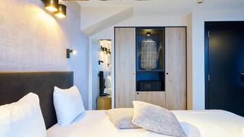 A modern hotel room with a large bed, a wooden headboard, a built-in shelf with decorative items, and a dark door.