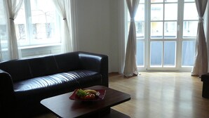 A living room with a black leather sofa, a wooden coffee table with fruit, and large windows with curtains. - FUKAS Apartments MEDENA (Bratislava)