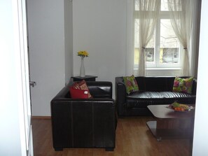 Un salon avec deux canapés en cuir noir, dont l’un est agrémenté d’un coussin rouge et l’autre de coussins aux couleurs vives. Un petit vase blanc contenant des fleurs jaunes est posé sur une table d’appoint.