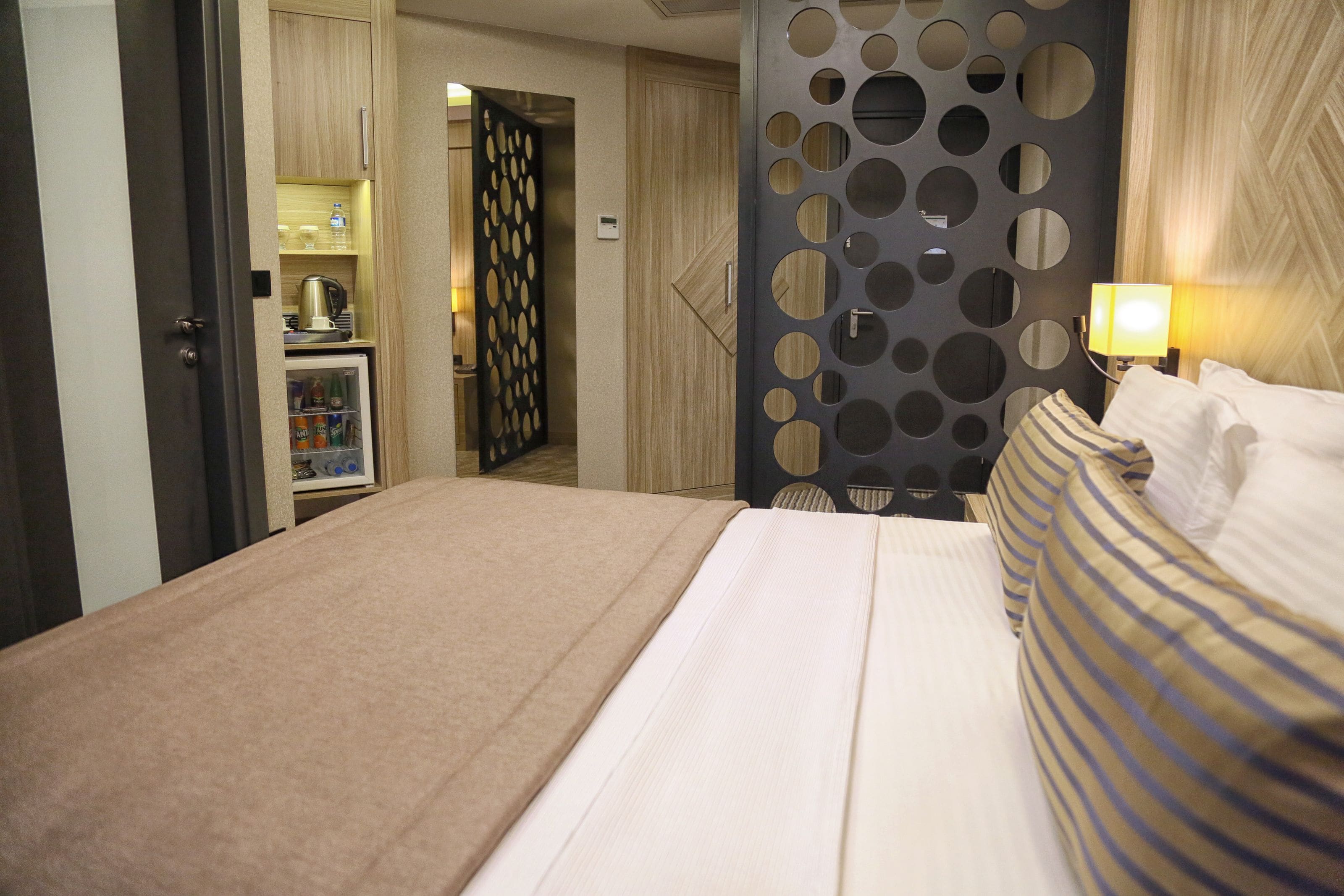 A hotel room with a bed, a minibar, a wall with circular cutouts, and a lamp.