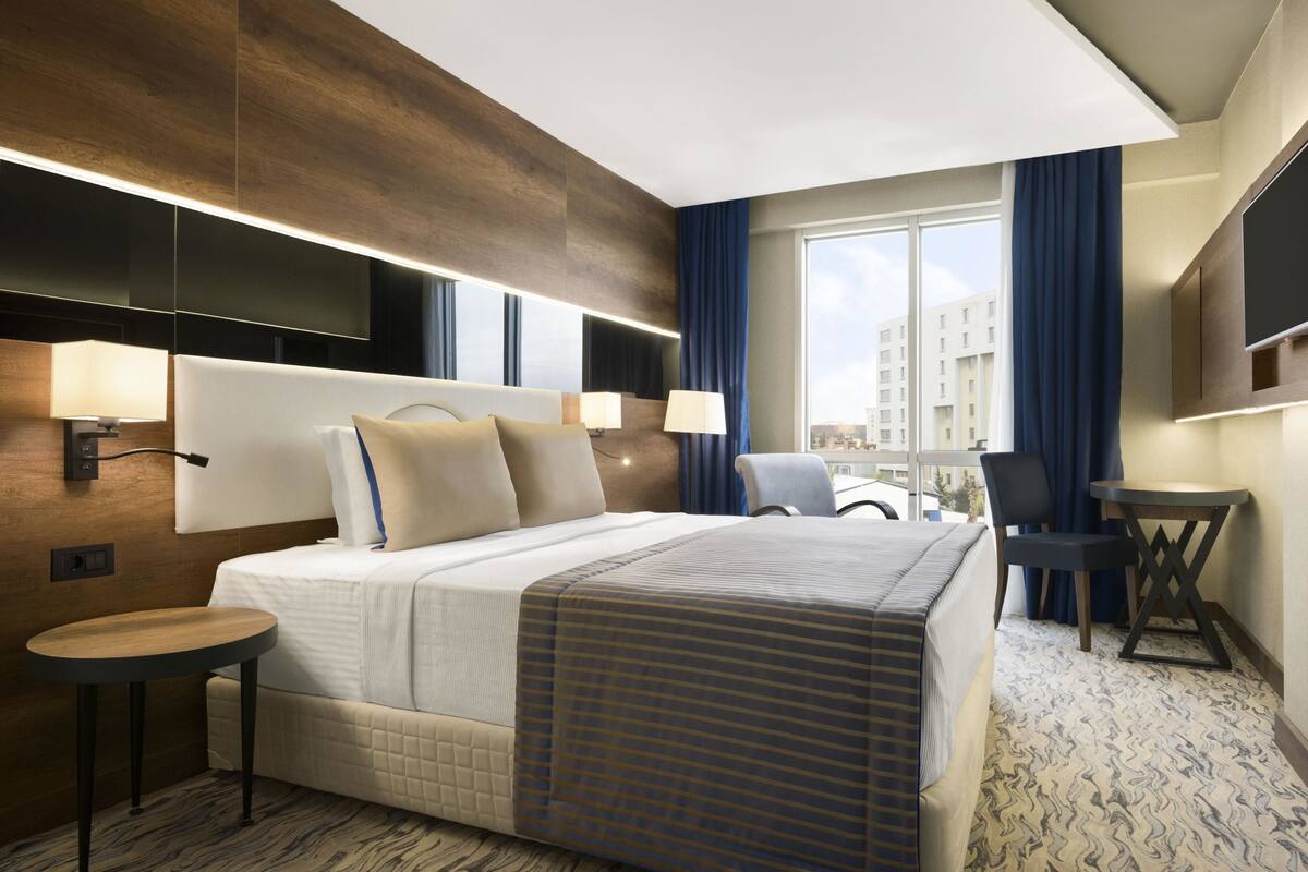 Een moderne hotelkamer met een groot bed, nachtlampjes, een klein tafeltje, een stoel, een televisie en een groot raam met gordijnen.