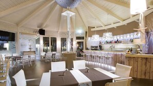 A restaurant with a high ceiling, white chairs, and tables with brown tablecloths. There is a bar area with bottles and a hanging light fixture.
