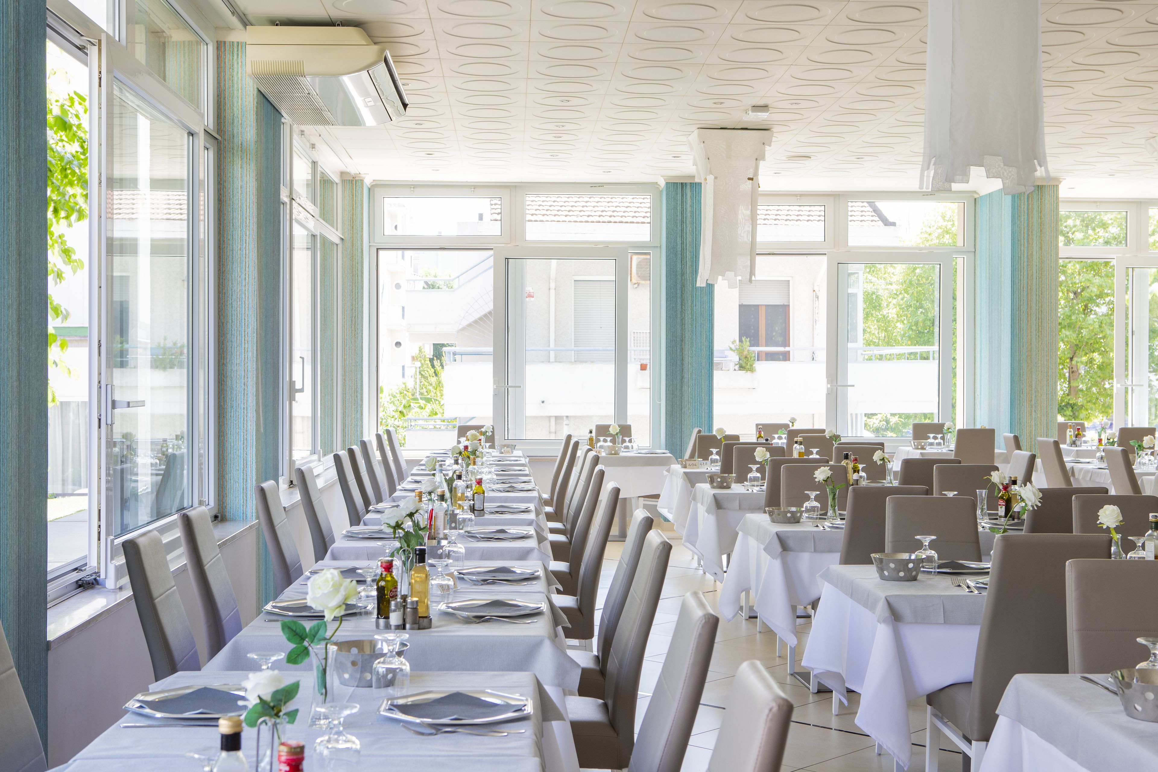 Un ristorante con lunghe tavole apparecchiate, ampie finestre e un soffitto con un motivo geometrico.