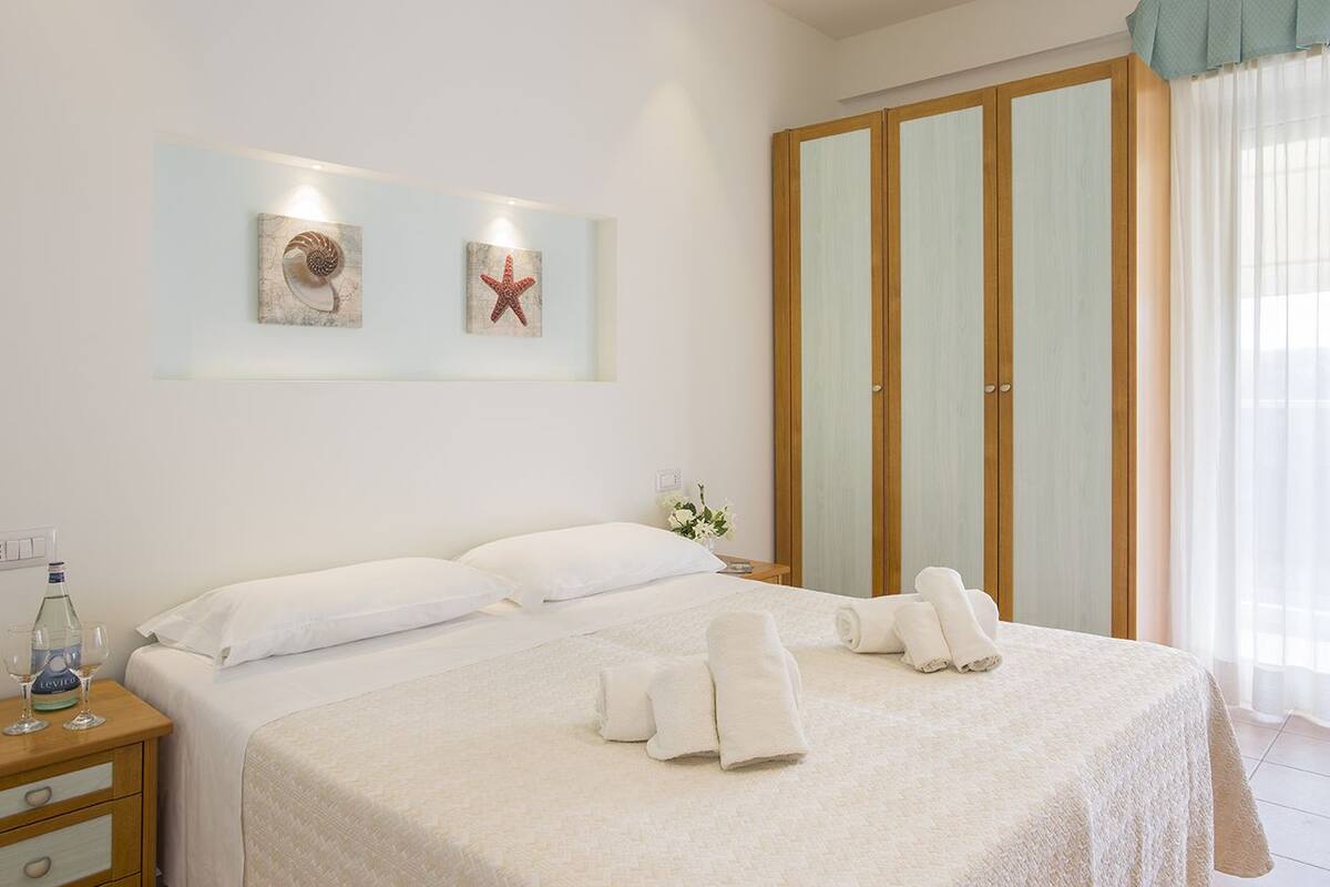 A neatly made bed with white linens and towels, a bedside table with a bottle and glasses, and artwork on the wall.