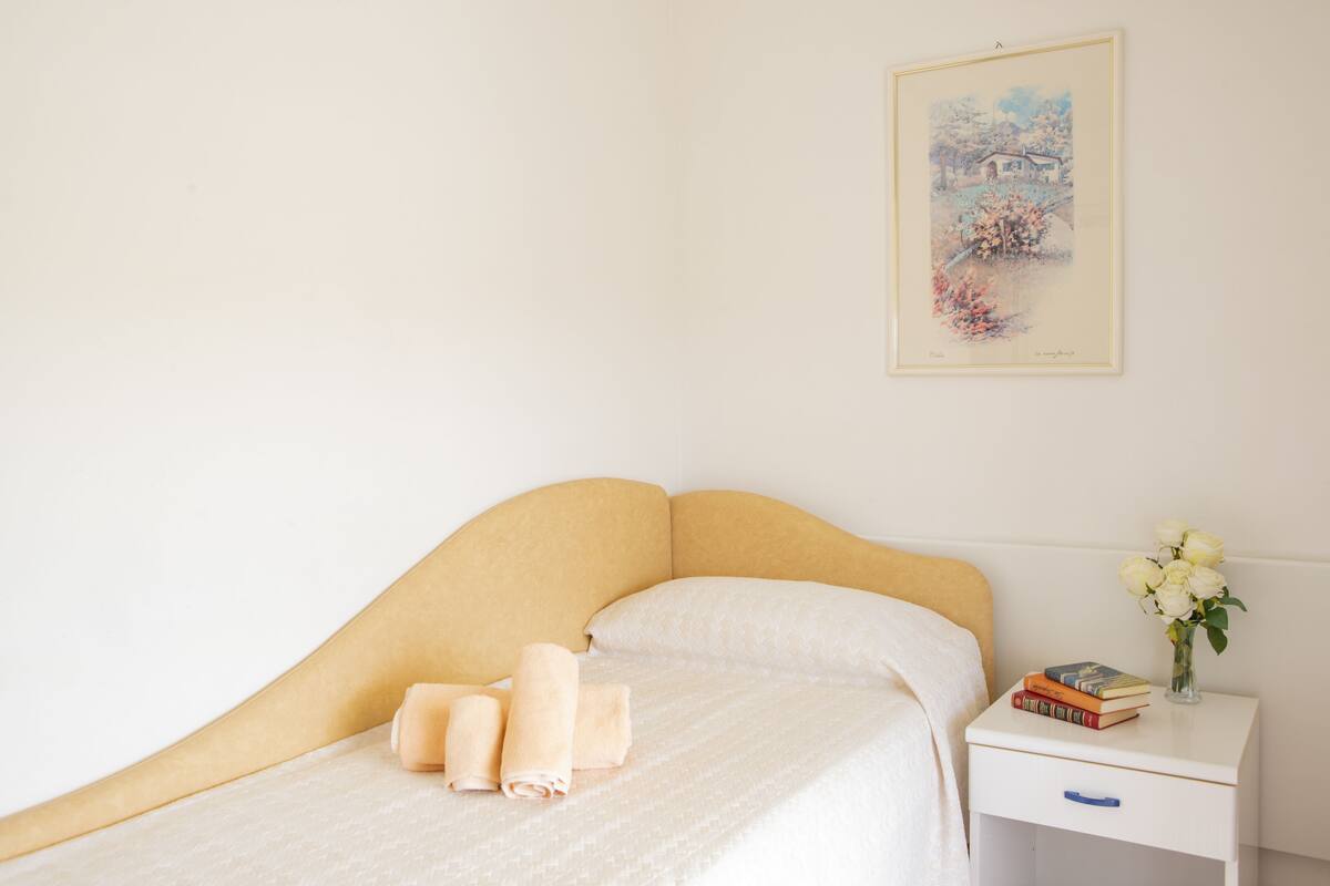 A neatly made bed with rolled towels, a bedside table with a vase of flowers, and a framed picture on the wall.