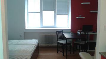A small room with a bed, a desk, and a chair. There is a window with blinds, a radiator, and a television on the desk.