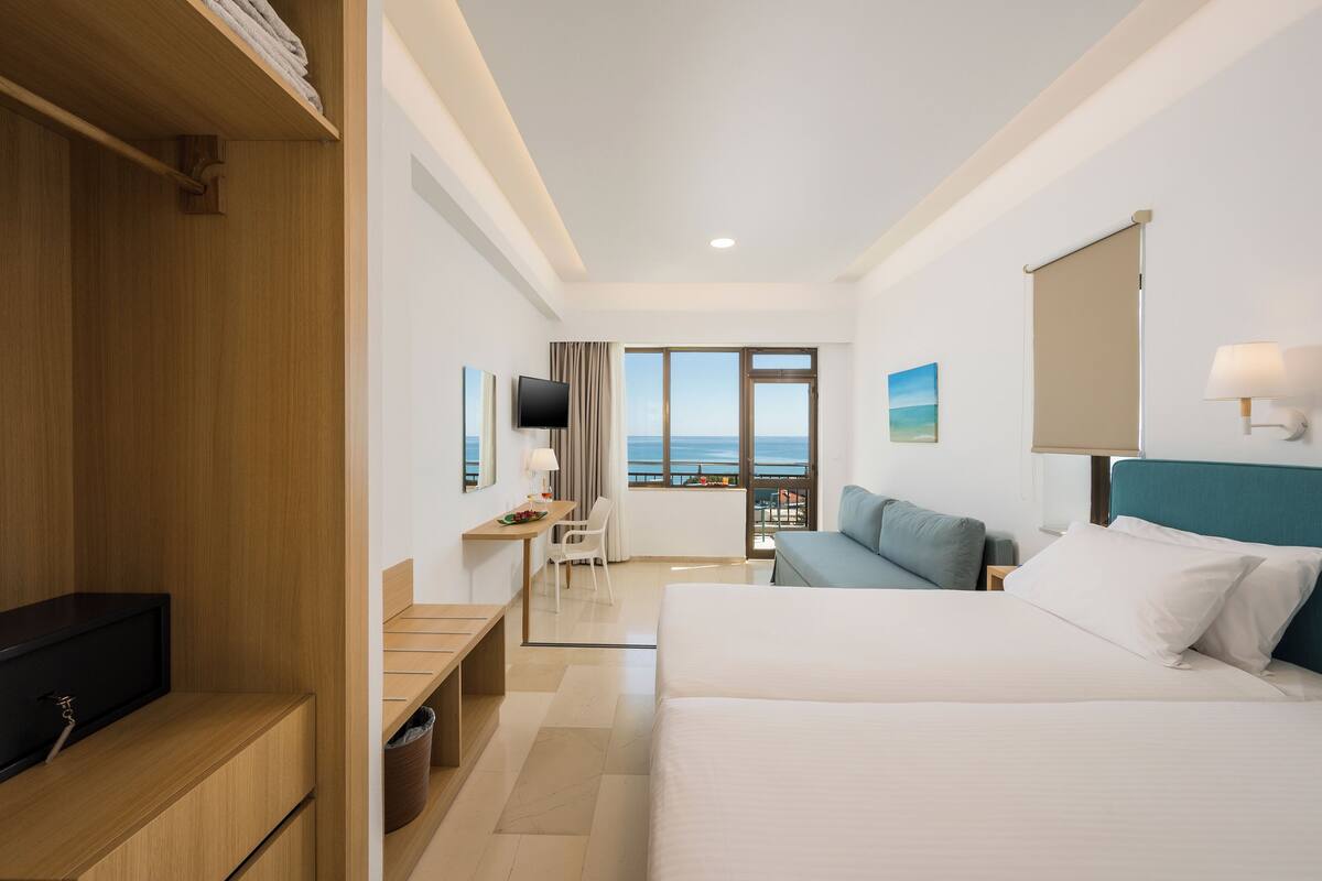 A hotel room with a bed, a desk, a TV, and a view of the sea.