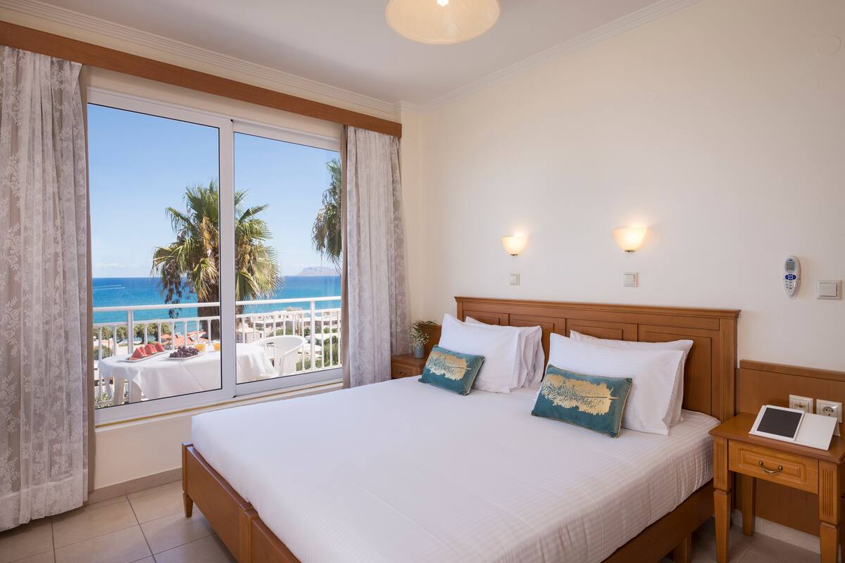 A hotel room with a large bed, a bedside table with a tablet, and a view of the sea and palm trees.