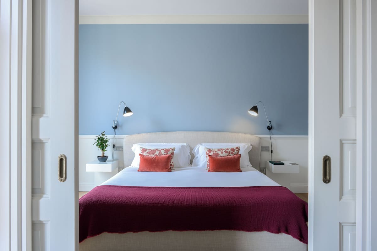 A bedroom with a bed, two bedside lamps, a nightstand with a plant, and a red throw blanket.