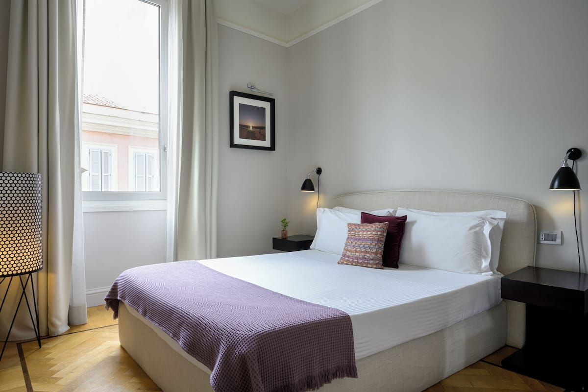 A neatly made bed with a white comforter and a purple throw blanket, a bedside table with a lamp, and a framed picture on the wall.