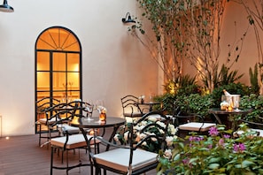 Un espace extérieur aménagé pour les repas, avec des tables et des chaises, une grande fenêtre en arc de cercle et des plantes en pot.