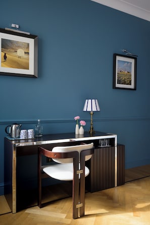 A room with a dark blue wall, a framed picture, a desk with a lamp, a chair, and a small cabinet.