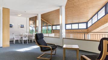 A room with a high wooden ceiling, a dining area with chairs and a table, and a large window with a view of a green landscape.