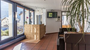 Uma área de recepção com balcão de madeira, grandes janelas, um vaso de planta e uma televisão montada.