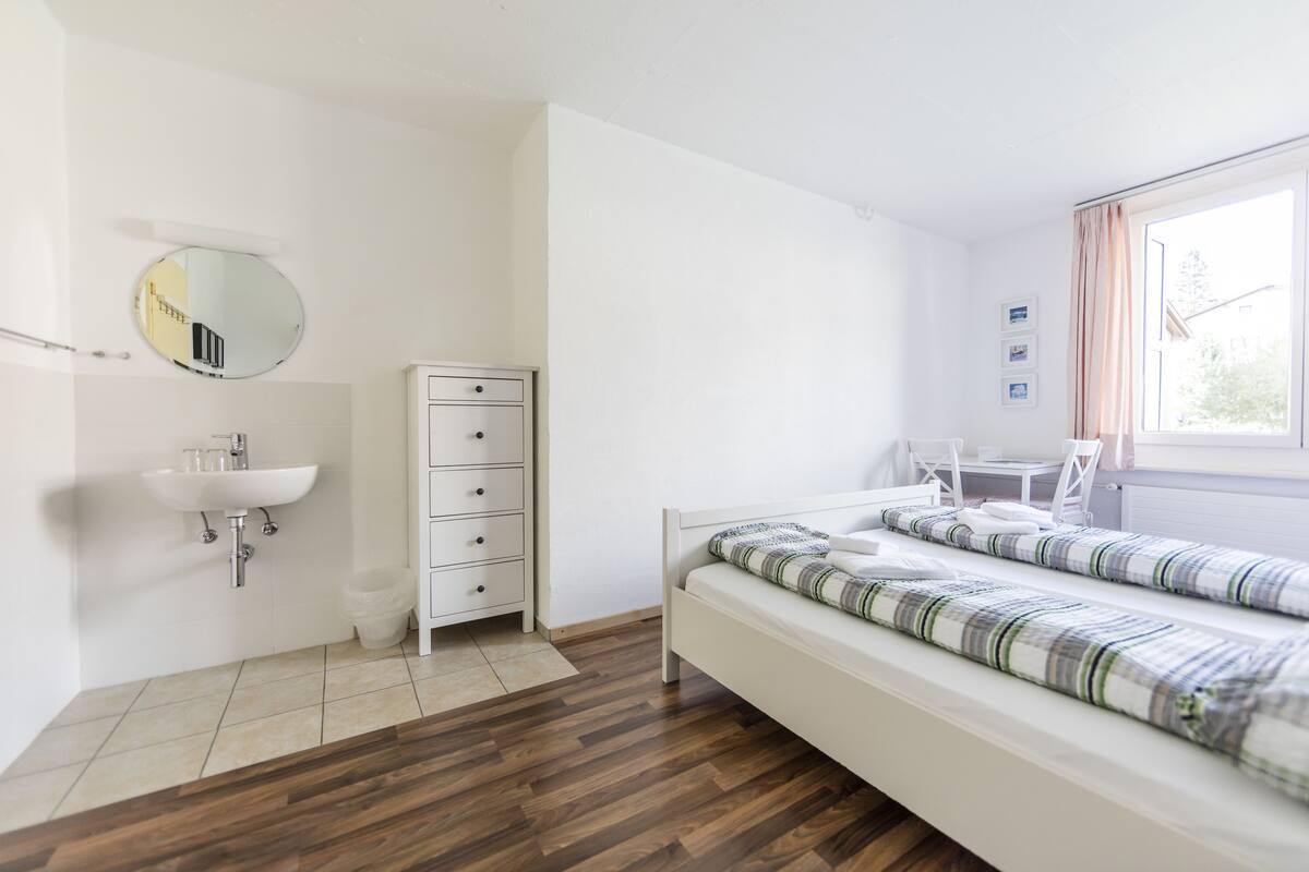 A bedroom with a wooden floor, a white bed with striped bedding, a white dresser, a round mirror, and a window with curtains.