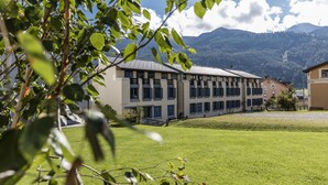 Um edifício com várias janelas e um telhado plano, rodeado por um espaço gramado e montanhas ao fundo.