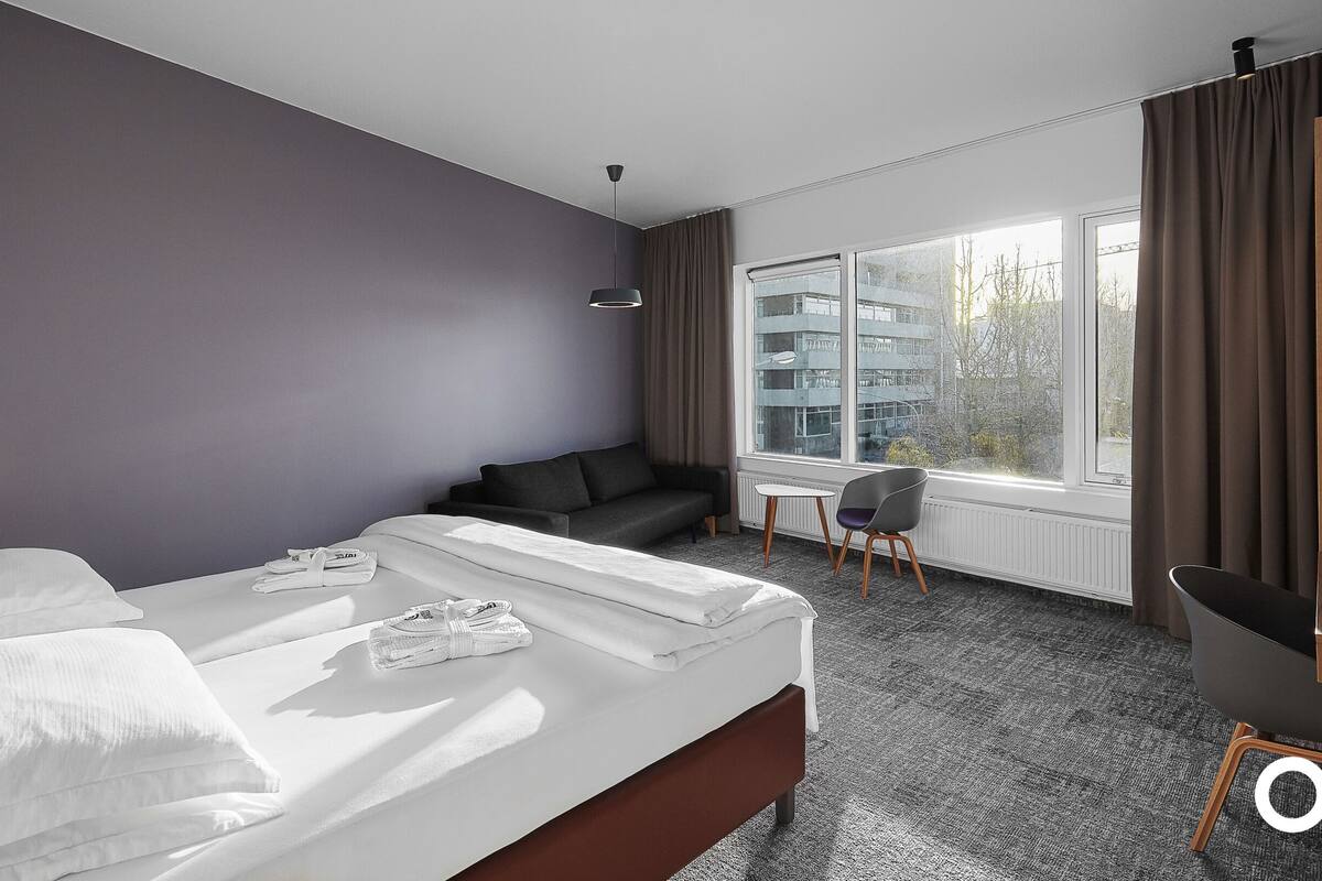 Et moderne hotelværelse med seng, sofa, skrivebord og stol. Der er et vindue med udsigt til bygninger.