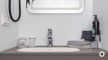 A modern bathroom with a white sink, a chrome faucet, and a mirror above it. There are two glass jars on the counter, a black bottle of shampoo, and a towel with "CENTURY HOTEL" on it.