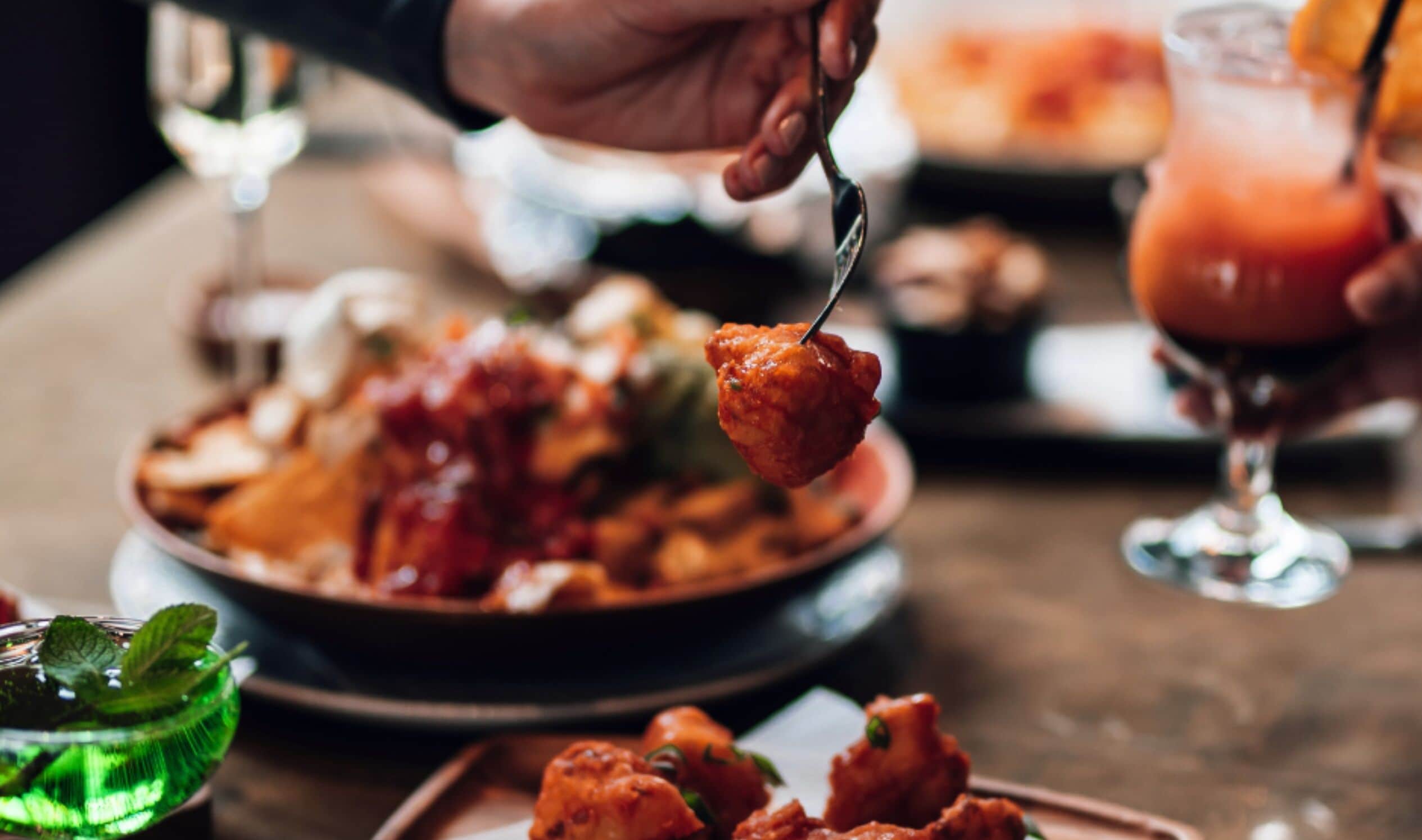 A person holding a fork with food, a glass of drink, and a plate of food on a table.